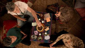 Still from the show Grace and Frankie. Overhead image of four people sitting at a table that has cups, paper, and various other objects sitting on it.